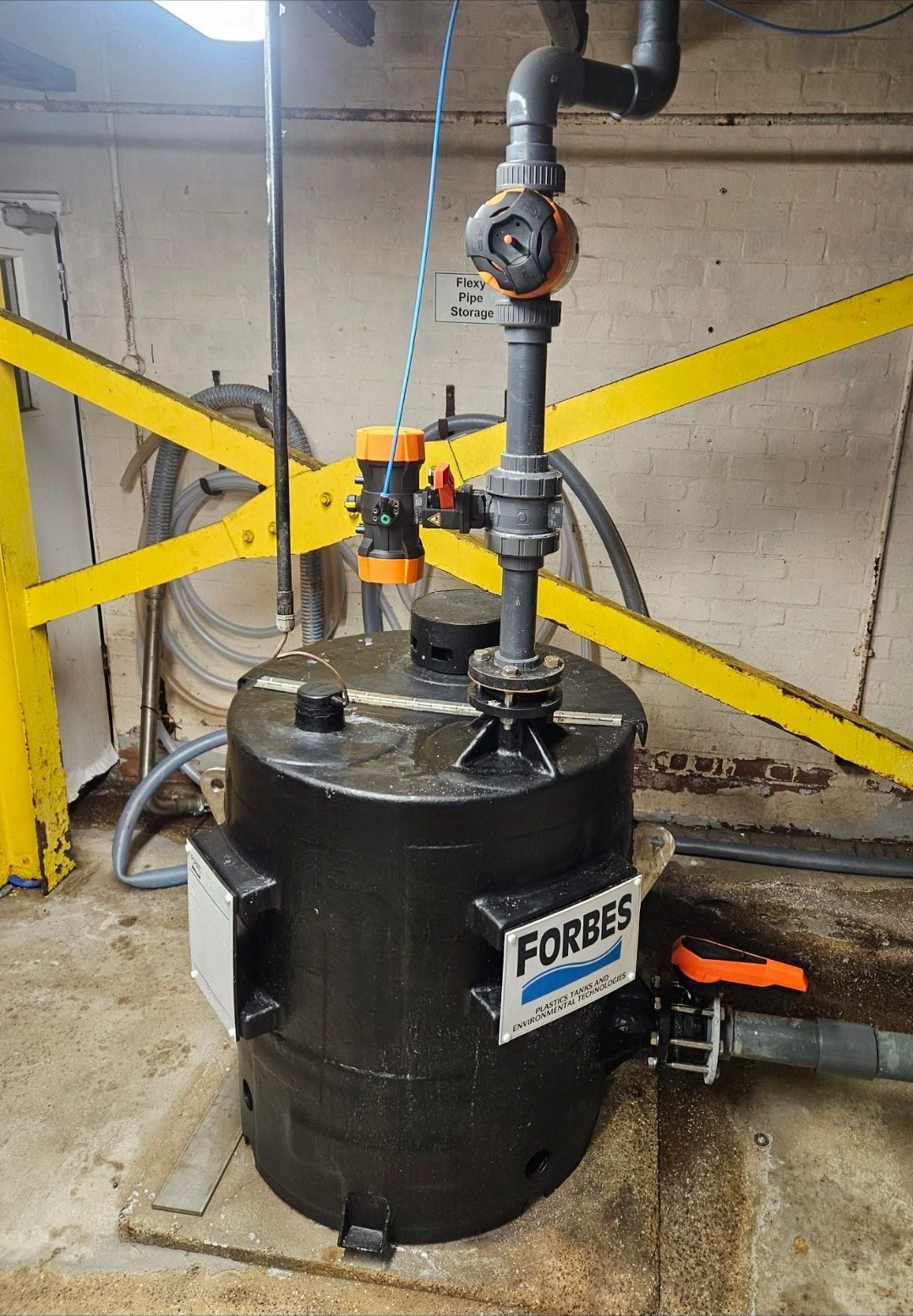 Black tank with new pipework and valves behind yellow safety barriers